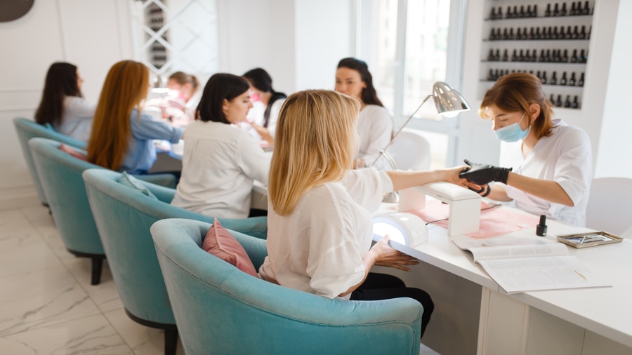 A nail technician wearing a mask and gloves giving a manicure to a client in a busy nail salon.