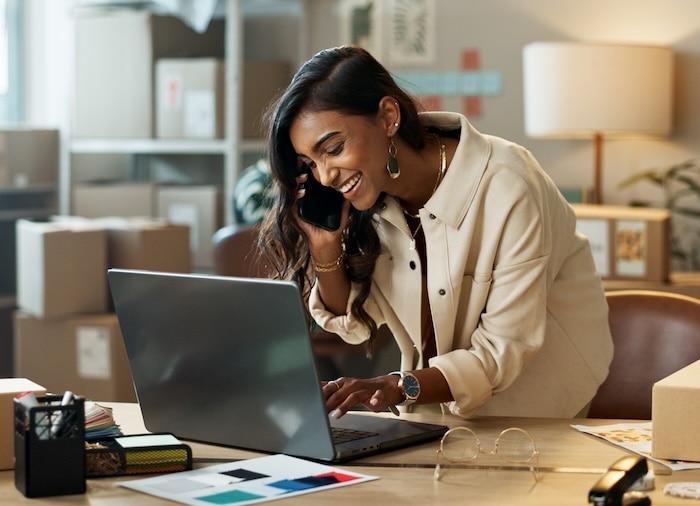 Smiling woman leaning over desk while talking on phone and typing on laptop.