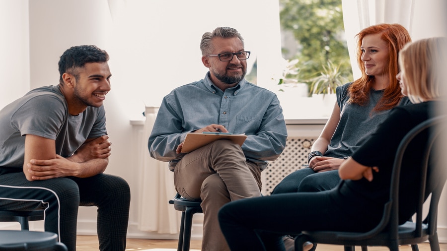 Four adults seated in a circle during a group discussion, with one person holding a clipboard and taking notes.
