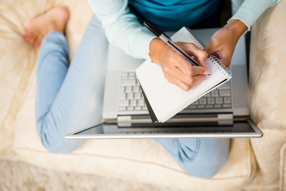 Woman sitting on couch with laptop open on her lap and notebook open on top of it taking notes