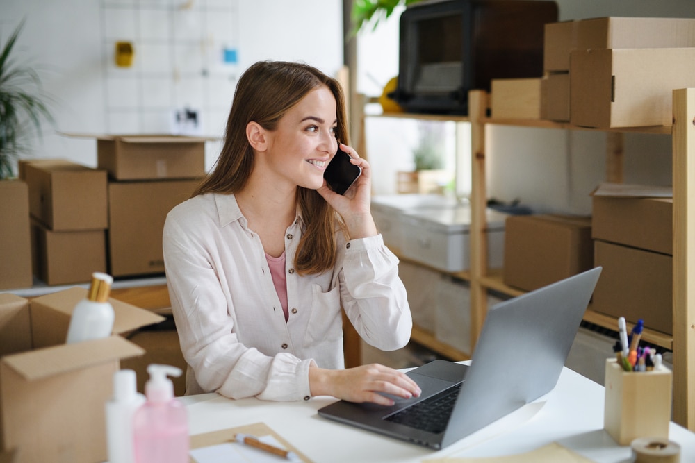 Woman happily smiling at a work desk in front of her open laptop while talking on the phone surrounded by boxes.