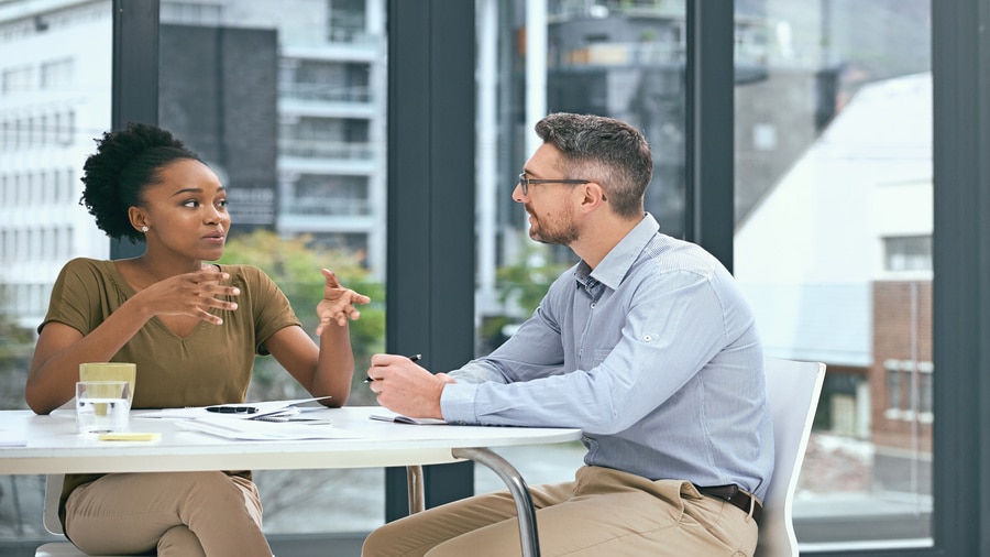 Two coworkers having a conversation at a table in a bright office, with papers and notebooks in front of them.