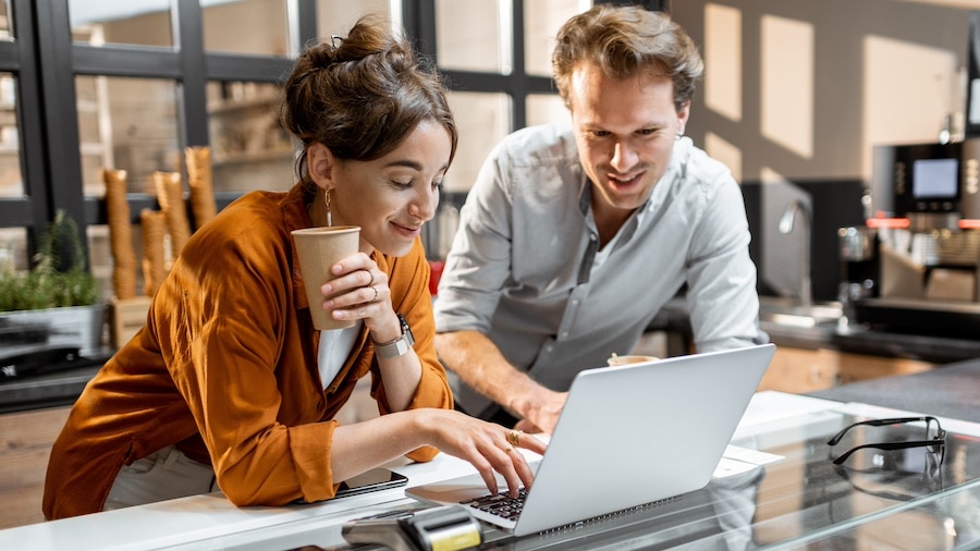 Two smiling people leaning over a desk and looking at a laptop.