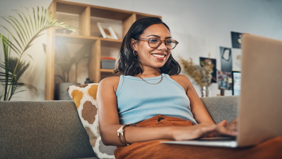 A woman smiling while working on a laptop from a couch in a home office.
