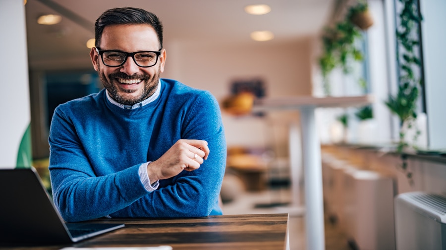 A smiling man wearing glasses and a blue sweater seated at a desk with a laptop in a modern office.
