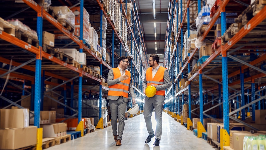Two people in orange vests standing in a warehouse, amongst the inventory.