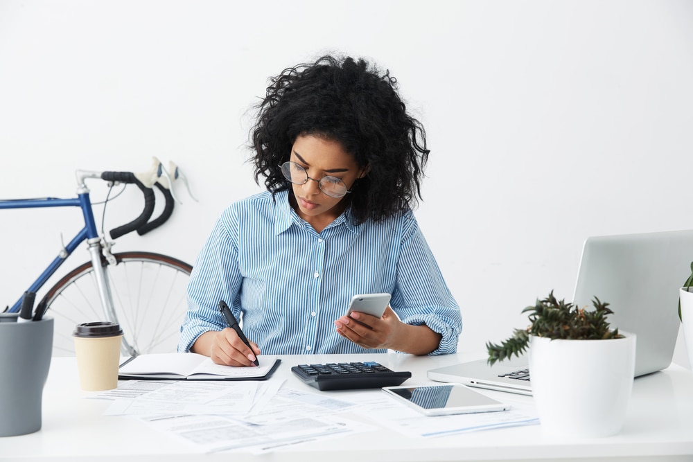 Young business woman writes in journal while looking at mobile phone