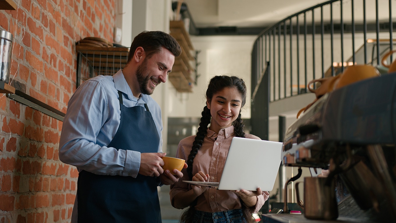 Two colleagues standing in a cafe reading about what's new in QuickBooks Online from a laptop.