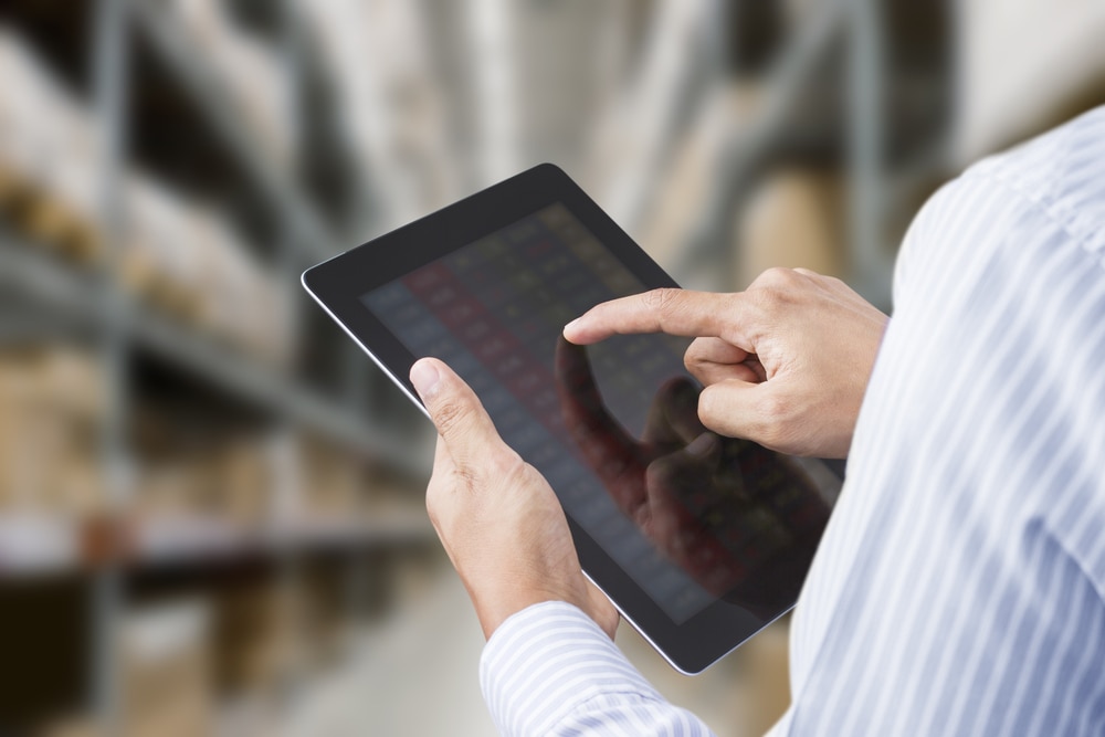 Man standing in a warehouse referencing a list on the tablet he's holding.