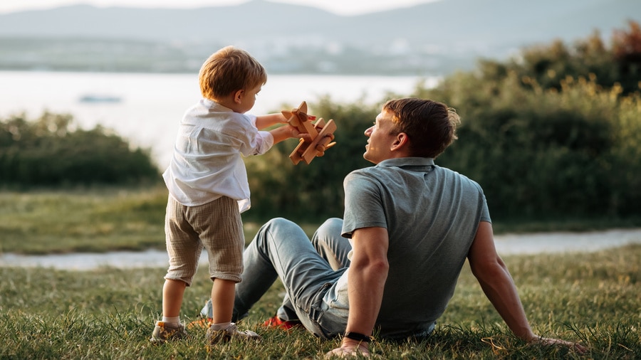 A father and young child playing with a toy airplane outdoors on a grassy hill near a lake.