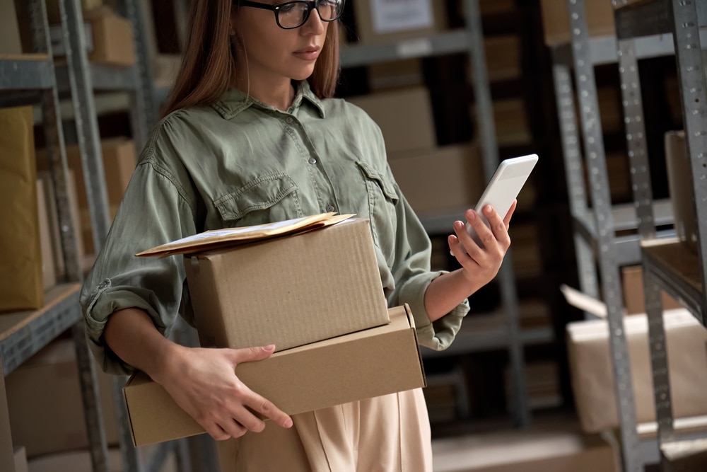Woman standing in a stock room, holding several packages under her arm while reading from her phone