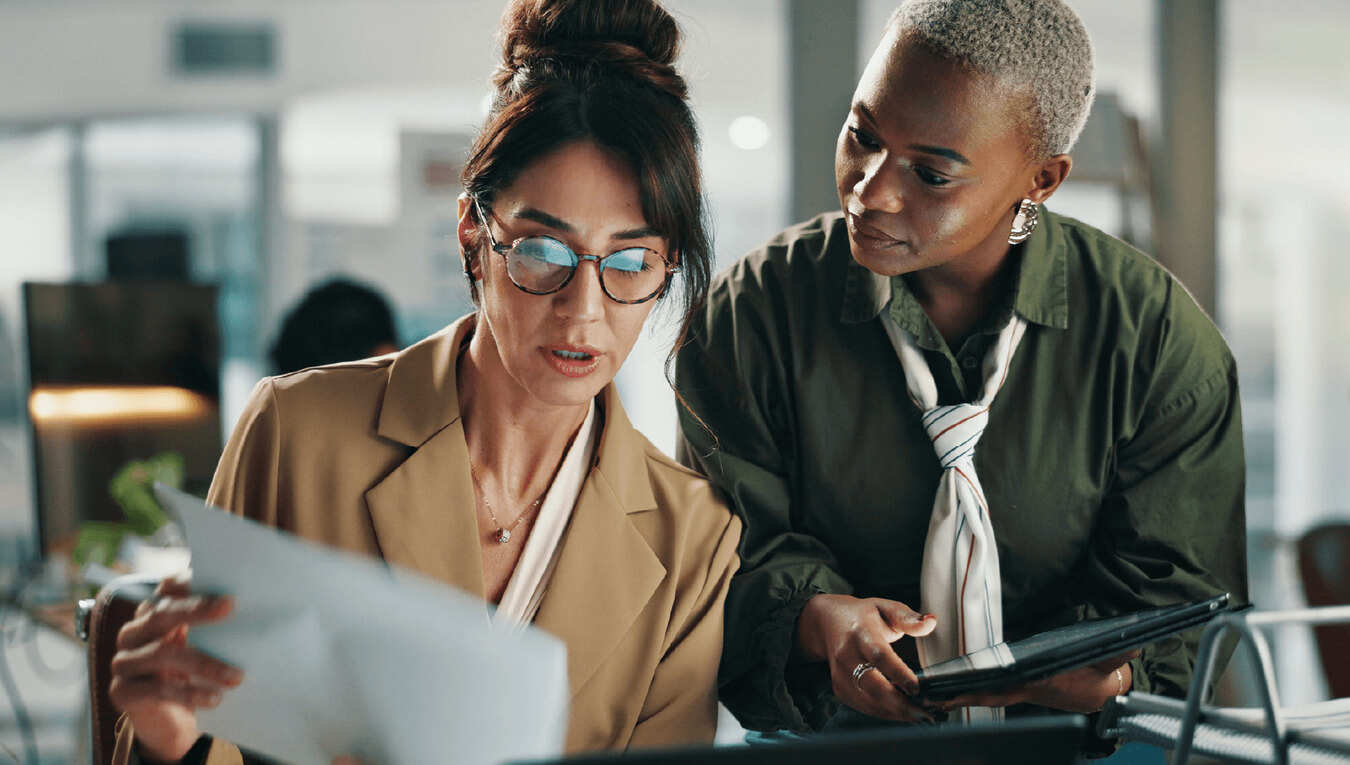 Two finance professionals reviewing a printed document together at a desk, one holding a tablet, discussing the numbers.
