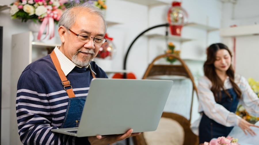 Two people in a flowershop. One looking at laptop in the foreground while the other wraps flowers in the background.