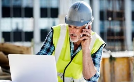 A photo of a construction worker wearing a hard hat, on the computer and on his phone.