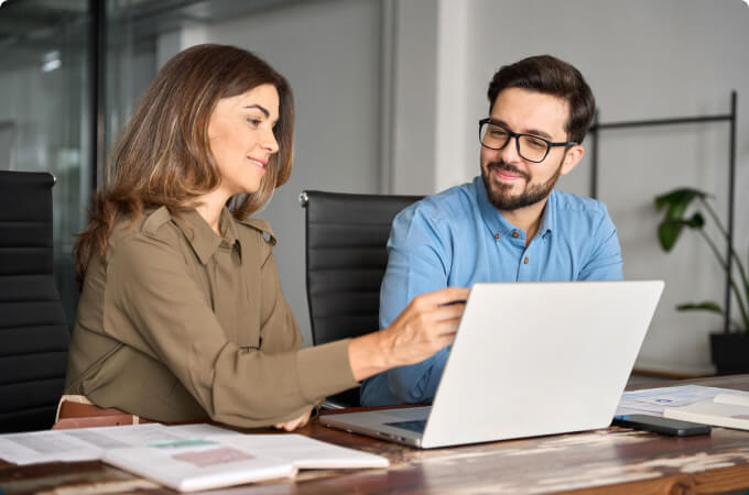 two people sitting at a table with laptop while pointing at something on screen