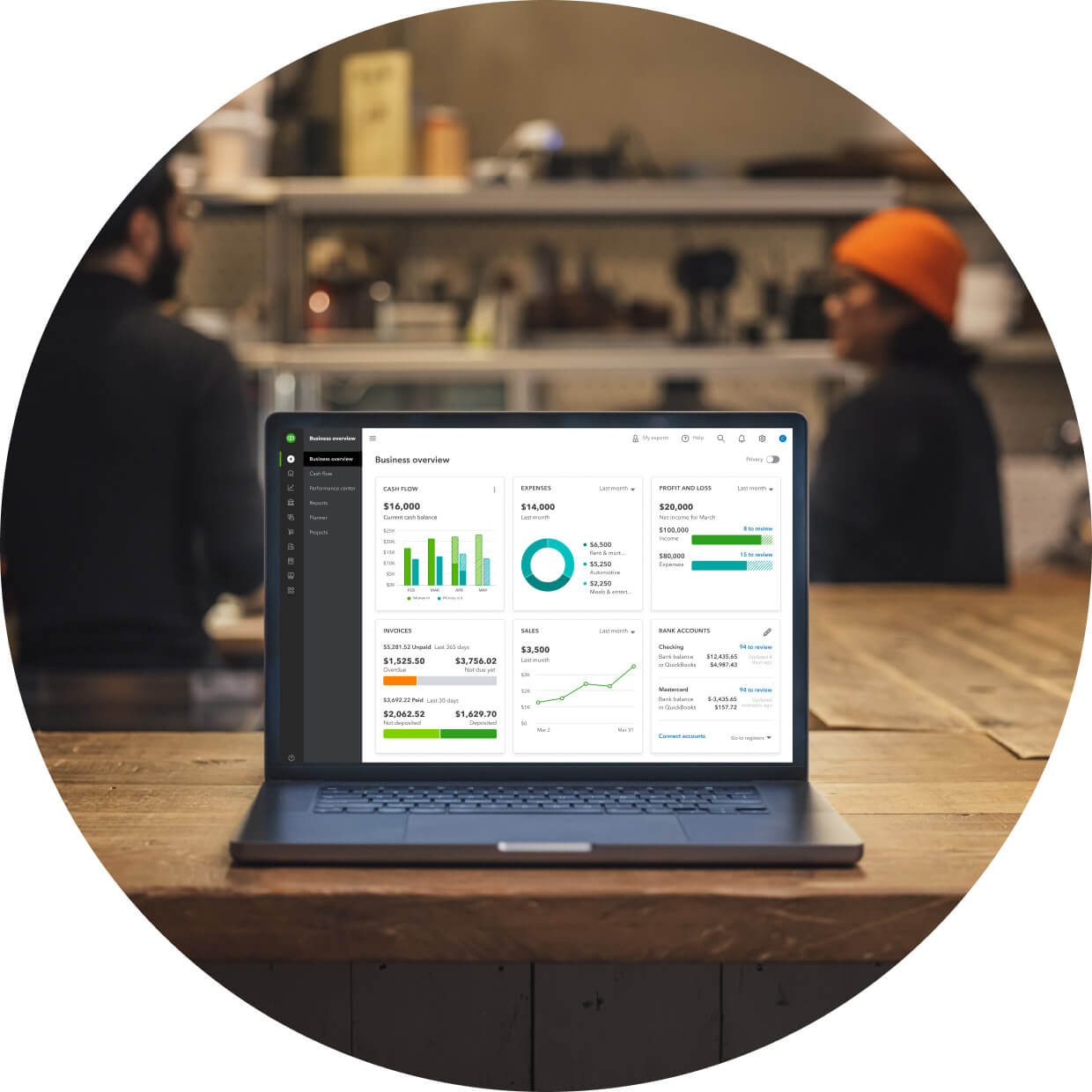 laptop open to QuickBooks dashboard on a table with two people in the distance