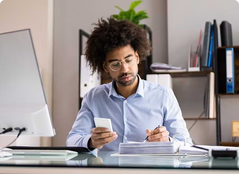 A person sitting at a desk is holding a mobile device in one hand and writing with a pen with their other hand. 