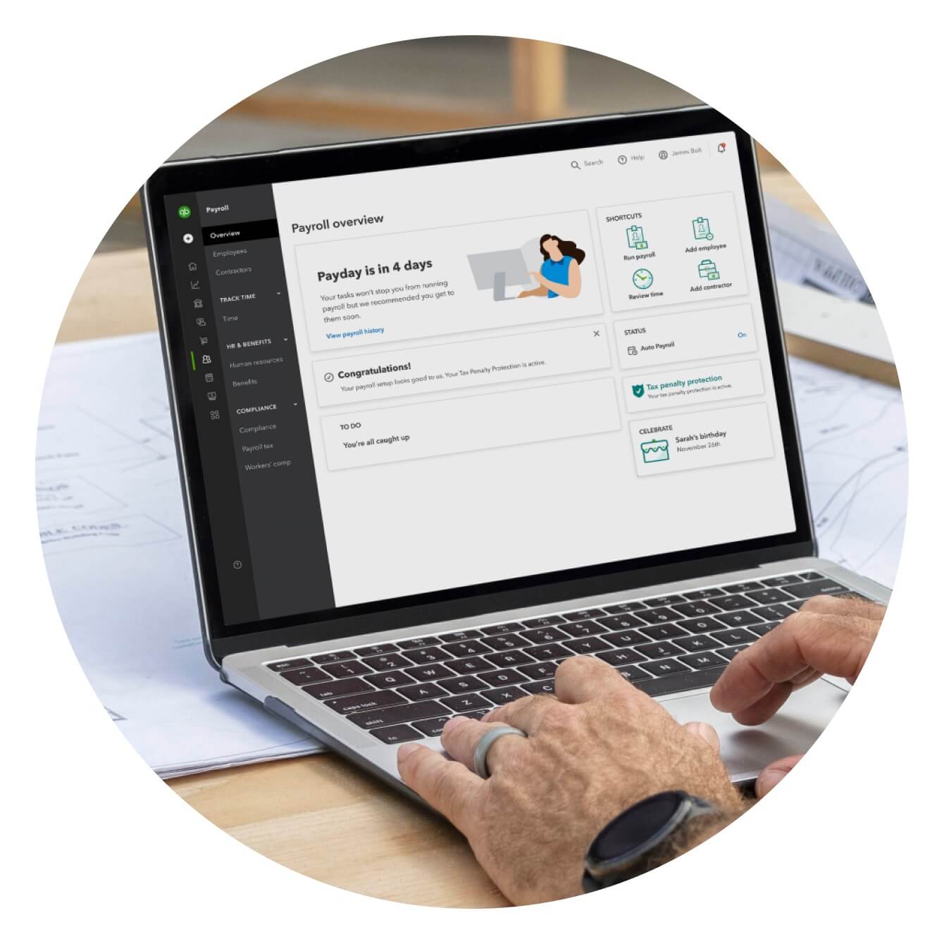 A photo of a laptop displaying the Payroll overview page in the QuickBooks Online platform. The laptop is sitting on a table outside at a construction site with a person working on it.