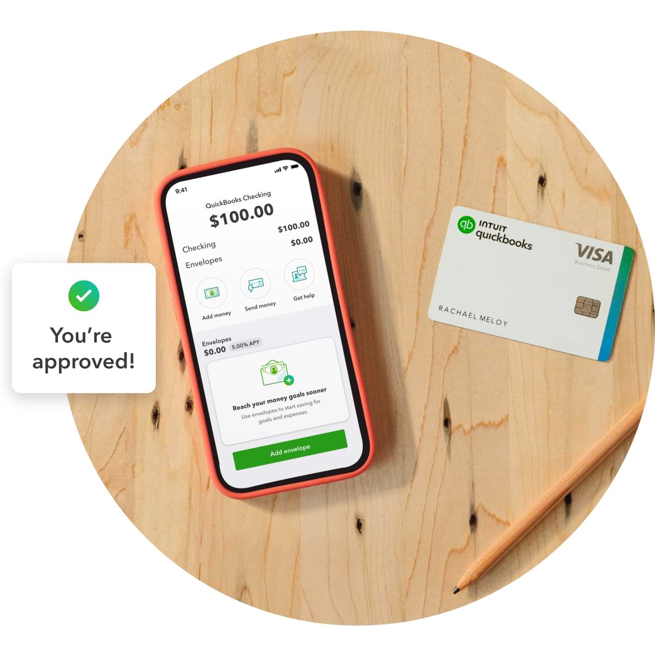 Desk with a QuickBooks debit card and a phone open to a business bank account displaying checking and envelope balances. A notification from QuickBooks reads “you’re approved!”