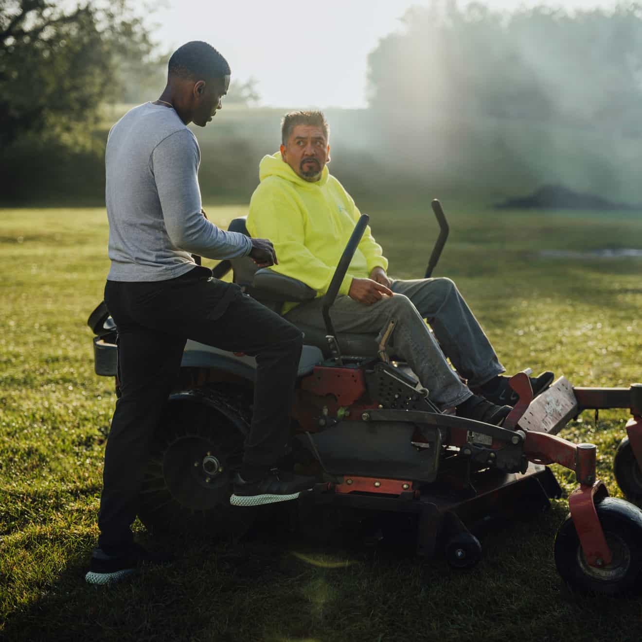 A landscaping business owner speaks to his employee who is operating a lawn mower.