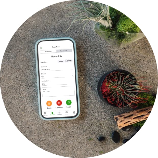 A mobile phone set on a concrete surface surrounded by plants, displaying the Track time screen of the Workforce app.