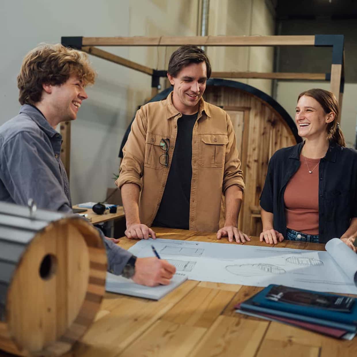 Three people are sitting around a drafting table, smiling and laughing together.