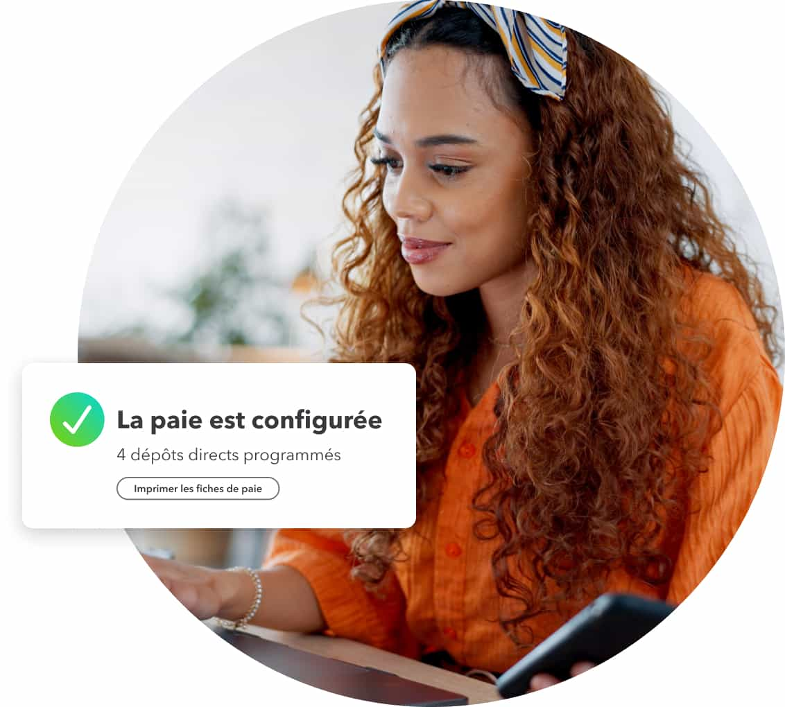 Une femme aux cheveux bouclés portant une chemise orange travaille sur son portable avec son téléphone montrant que la paie est configurée dans QuickBooks.