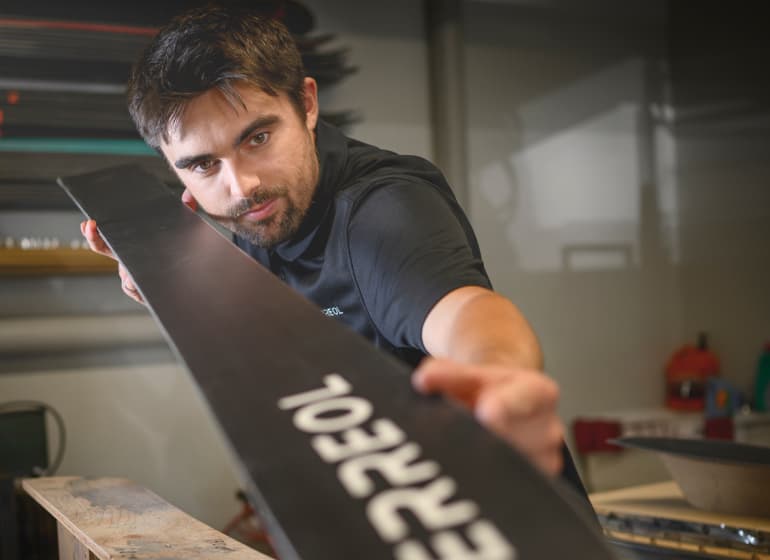 Un homme barbu examine attentivement la courbe d'un ski qu'il fabrique.