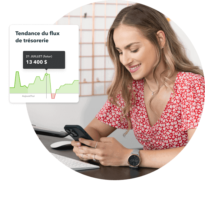 Une femme assise à son bureau vérifie les flux de trésorerie dans QuickBooks sur son smartphone