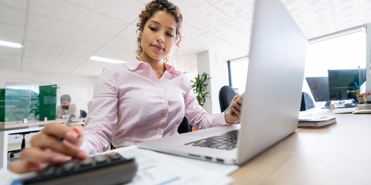 imagem de uma mulher usando camisa cor de rosa sentada em frente a um notebook e utilizando uma calculadora ilustrando artigo sobre dva na contabilidade